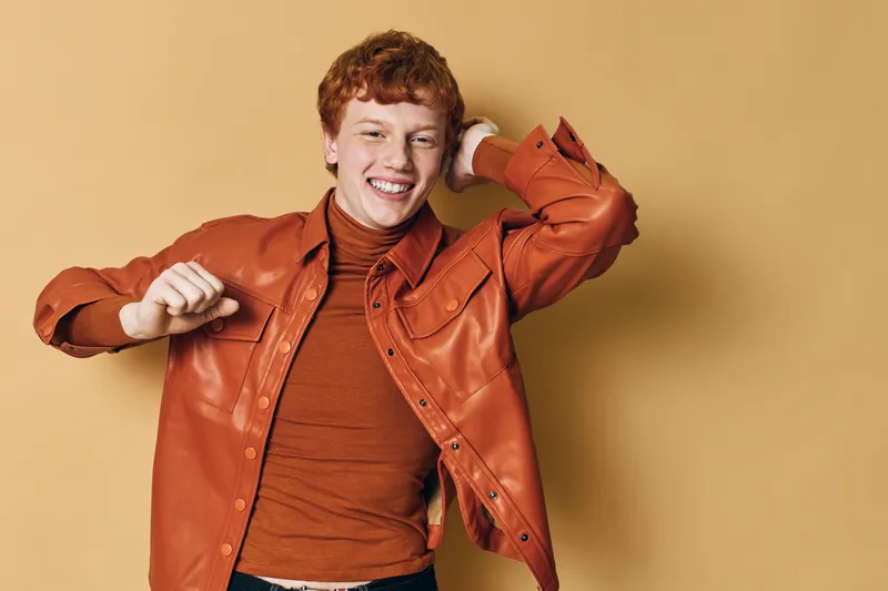 Young man with curly red hair wearing brown leather jacket and turtleneck smiling and dancing on beige background with happy expression and casual style.