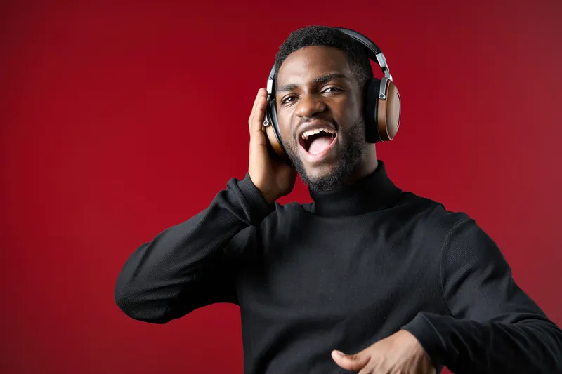 Joyful young Black man enjoying music with headphones on a vibrant red background, expressing happiness and energy through singing and dancing