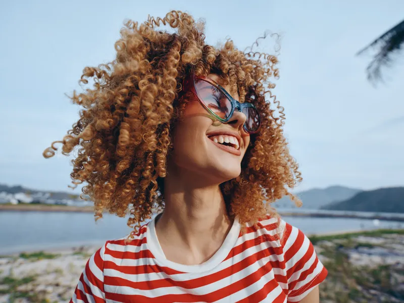 A smiling woman with voluminous curly hair wearing stylish sunglasses and a red and white striped shirt, captured outdoors against a bright seaside landscape and blue sky.