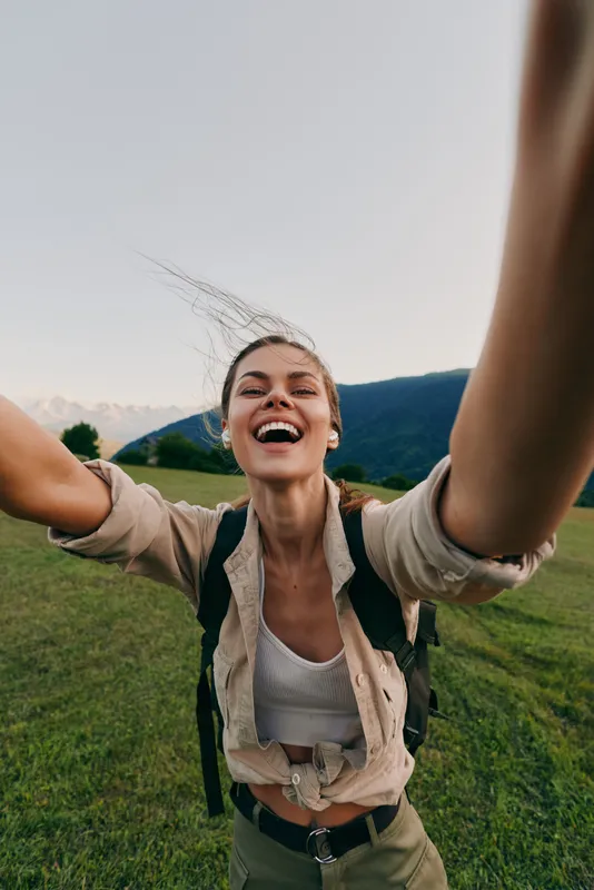 Young woman with backpack enjoys a carefree outdoor moment, smiling broadly as she reaches toward the camera in a grassy field with distant mountains.