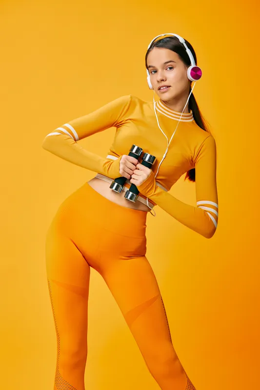 Young woman in bright orange fitness outfit holding small dumbbells, wearing pink headphones and posing confidently for a studio fitness portrait on a warm orange background.