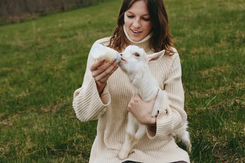 A young woman in a beige sweater feeds a baby goat with a bottle while sitting on grass in a natural outdoor setting during a lifestyle moment.