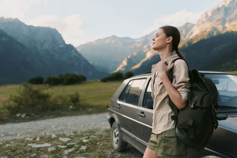 Young woman with a backpack stands beside a car in a mountain valley enjoying fresh air and peaceful nature during a hiking trip on a sunny day.