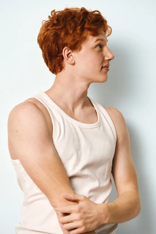 Redhead man with curly hair in white tank top standing and looking to side posing against white background with relaxed expression for casual style portrait.