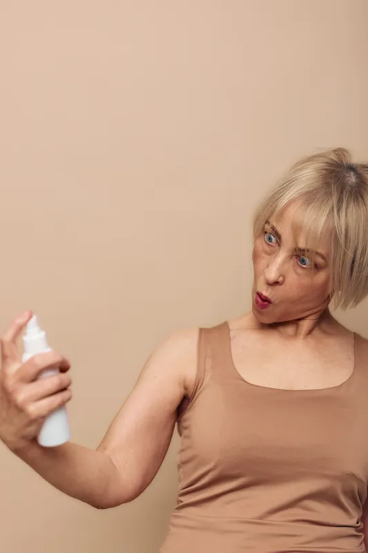 Surprised mature woman in beige tank top sprays deodorant on her arm against plain background. Concept of personal hygiene, skincare routine, and daily self-care habits.