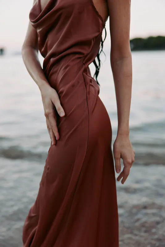Close crop of a burgundy satin dress on a slender figure by the shoreline, sunlight tracing soft folds and glossy fabric, creating an elegant, tranquil mood.