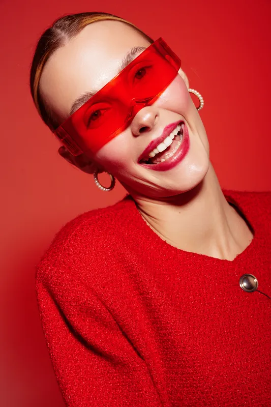 Smiling woman in red sweater and red transparent glasses poses against red background. Fashion portrait with bright makeup and joyful expression. Concept of style and confidence.
