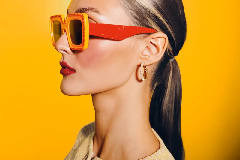 Profile of stylish woman with ponytail wearing large orange sunglasses and hoop earrings on bright yellow background. Concept of fashion, beauty, and summer accessories.