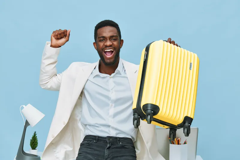 Vibrant image of a confident smiling man with dark skin holding a yellow suitcase in one hand, dressed in a white shirt and blazer, celebrating success at a modern workspace with blue background. Peop