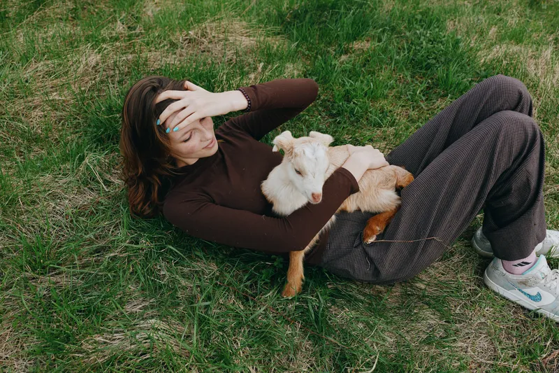 Relaxed woman lying on grass outdoors with a playful goat kid resting on her chest, sunlit moment, casual clothes, gentle breeze, natural colors, tranquil countryside vibe