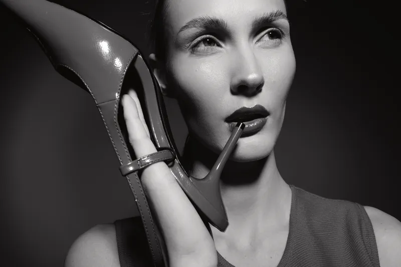 Caucasian woman with fair skin holds high heel shoe close to face in artistic black and white portrait with dramatic lighting and thoughtful expression.