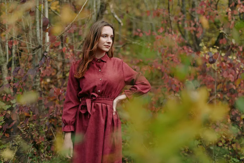 Genuine portrait of a young woman in a simple red dress standing among autumn foliage, conveying authentic natural presence, subtle emotion and a credible documentary style atmosphere.