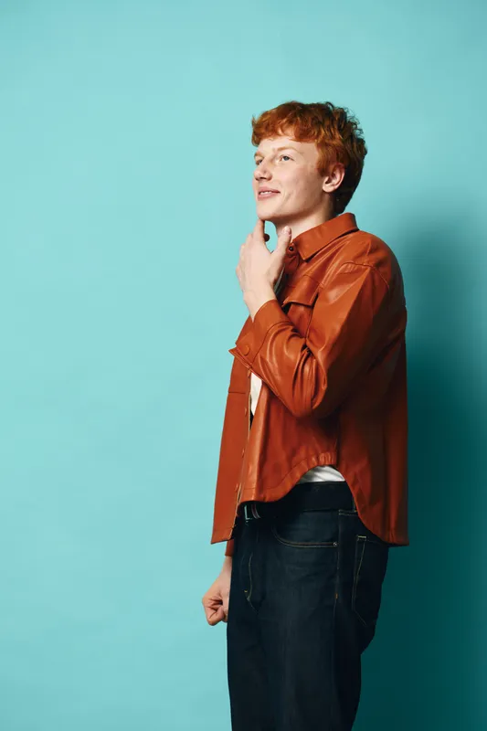 Young man with red hair wearing brown leather jacket and black jeans standing against blue background looking sideways with thoughtful expression and hand on chin.
