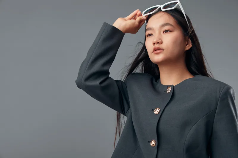 Young woman with long dark hair adjusting white sunglasses on head wearing dark gray coat posing on gray background in studio with confident facial expression and elegant style.