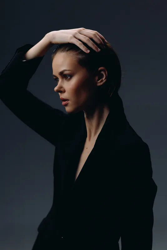 Thoughtful woman in black outfit posing with hand on head against dark background with moody lighting.