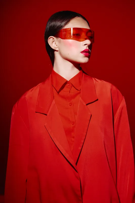 Young woman in a bright red oversized blazer and matching shirt poses confidently with glossy red lips, sleek hair and futuristic red translucent sunglasses on a solid red background.