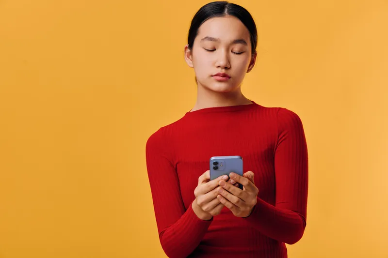 Young woman with long hair in a red sweater looking at smartphone with closed eyes on yellow background. Person using mobile device for communication or social media concept.