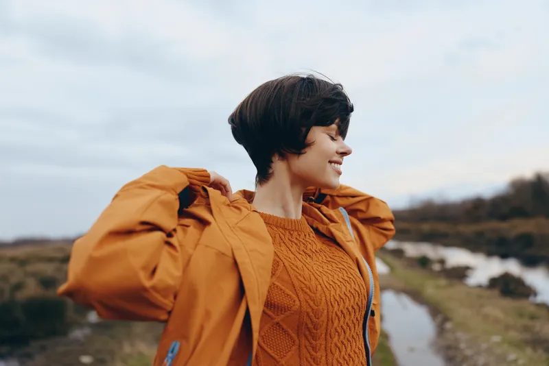 Woman lifestyle outdoors, orange jacket, coastal path by ocean, gentle smile, wind moving hair, nature scenery and travel feeling, cloudy sky background