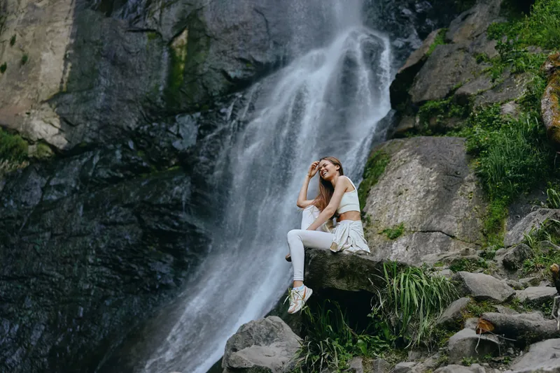 young woman sitting near waterfall wearing white activewear, enjoying nature outdoors with rocks and greenery around, serene and peaceful atmosphere