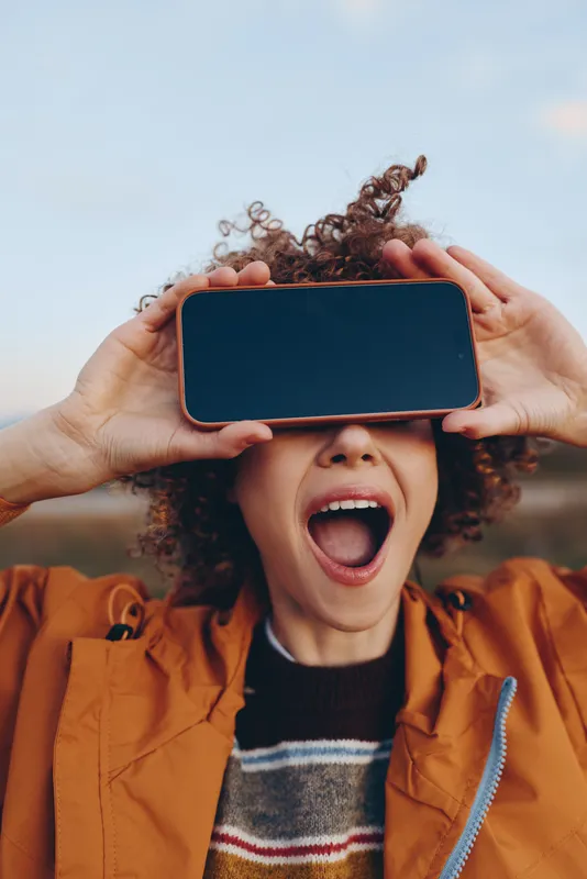 Woman lifestyle outdoors, curly hair in soft breeze, hands hold smartphone to cover eyes, laughing with open mouth, playful portrait, warm jacket, golden daylight, blurred background