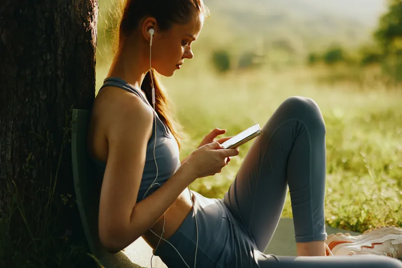 A young woman in sportswear enjoying music on her smartphone while sitting outdoors in a peaceful green landscape, portraying a relaxed and active lifestyle