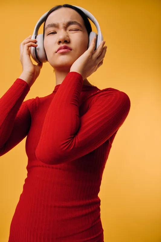 Woman in red sweater wearing headphones with closed eyes listening to music on yellow background. Girl enjoying sound and feeling relaxed with wireless audio device and style.