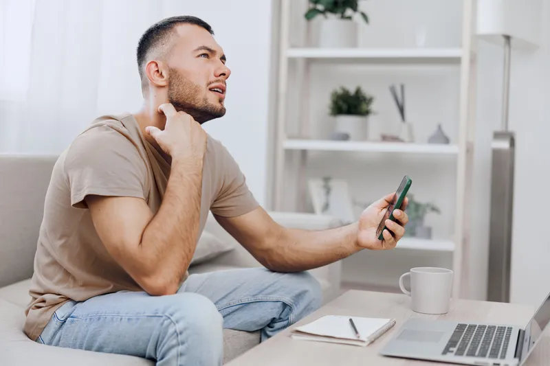 Thoughtful man sitting on a sofa, holding a smartphone and contemplating, showcasing modern lifestyle with a hint of creativity and relaxation.