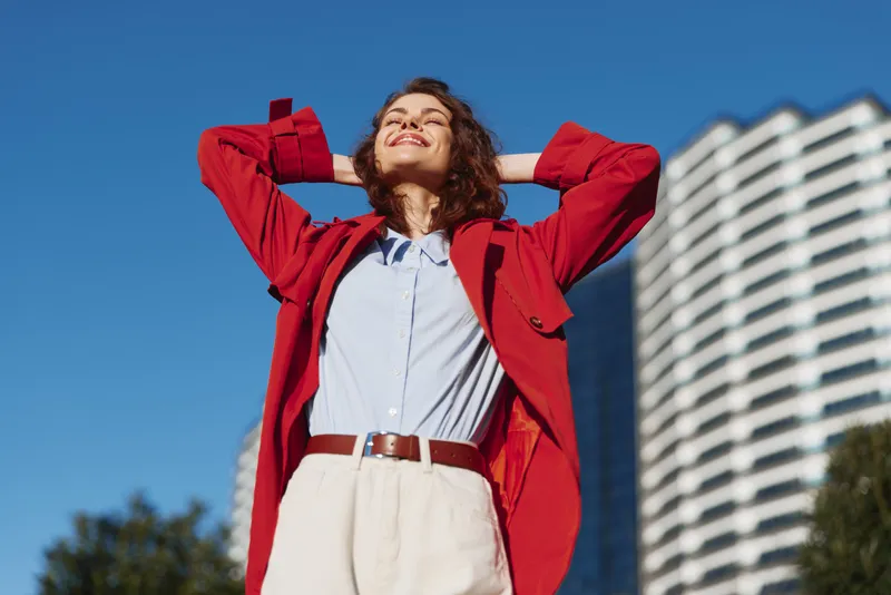 Young woman in red jacket stands outdoors with hands behind head smiling with closed eyes enjoying sunny day near modern building under clear blue sky.