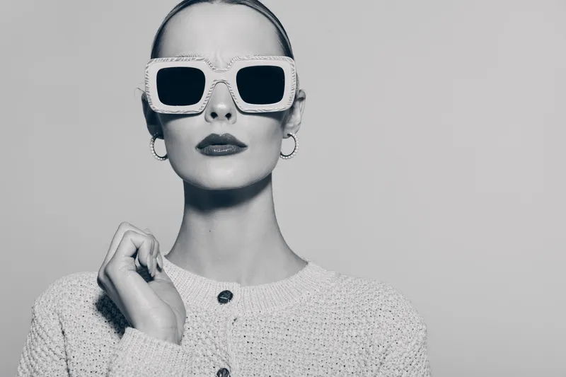 Elegant white woman with pale skin wearing oversized sunglasses and textured sweater poses in monochrome studio portrait.