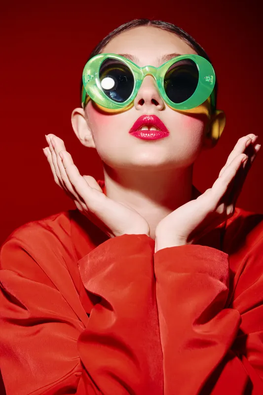 Young woman in bright green round sunglasses poses with red lips wearing vibrant red outfit and pink eyeshadow on red background. Fashion and beauty creative concept with intense colors.