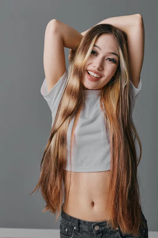 Young woman with long hair smiling and posing with raised arms, wearing a cropped t shirt and jeans in studio with grey background. Casual style, happy expression and natural look.