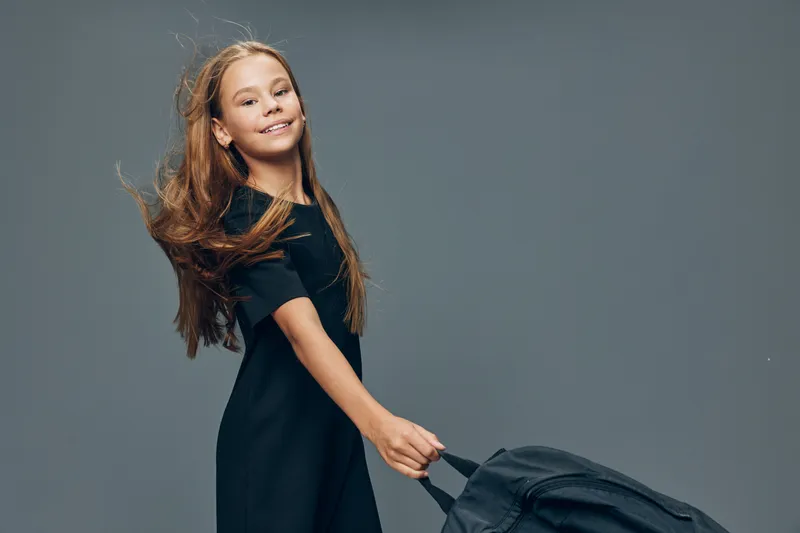 Girl, child and happy in black dress standing and holding suitcase with smile for travel, vacation or trip with wind in hair on grey background in studio.