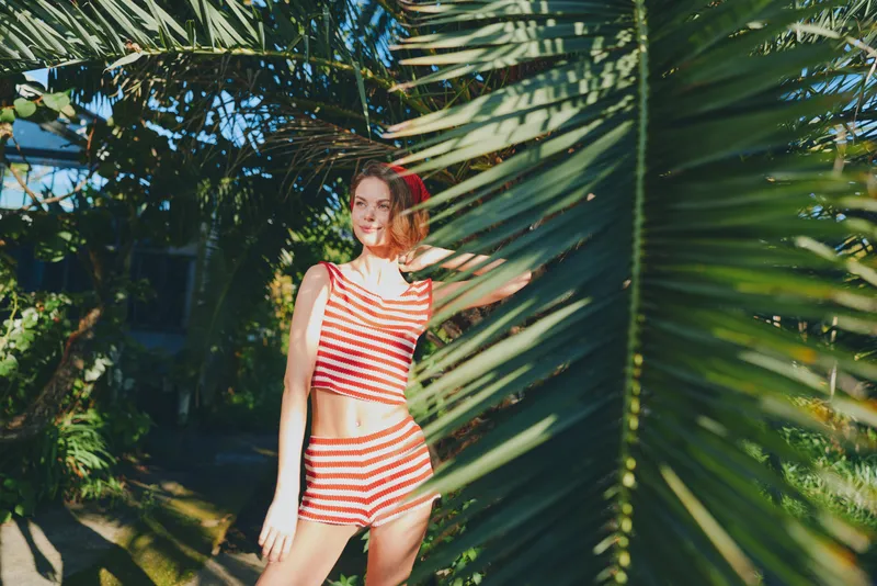 A cheerful woman stands among lush palm fronds in a sunny outdoor setting. She wears a red and white striped outfit, smiles brightly, exuding a vibrant, relaxed mood in a natural tropical environment.