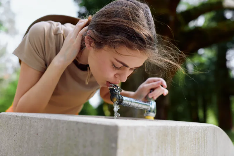 Woman drinking from a public fountain in a park, young female leaning over a stone basin to sip water, candid portrait of a refreshing hydration moment with green trees and sunlight
