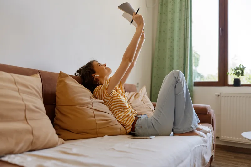A woman lounges on a couch at home, reading a book in warm sunlight, embodying lifestyle, relaxation, and casual comfort in a cozy interior setting. Her calm pose suggests quiet downtime.