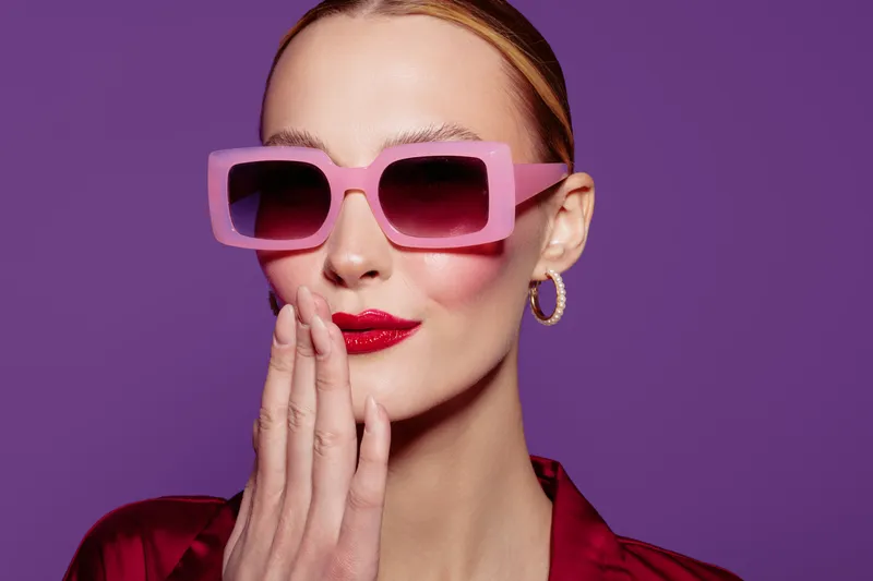 Stylish woman with pink sunglasses and red lipstick poses on purple background. Fashion portrait with elegant makeup and confident expression. Concept of glamour and style.