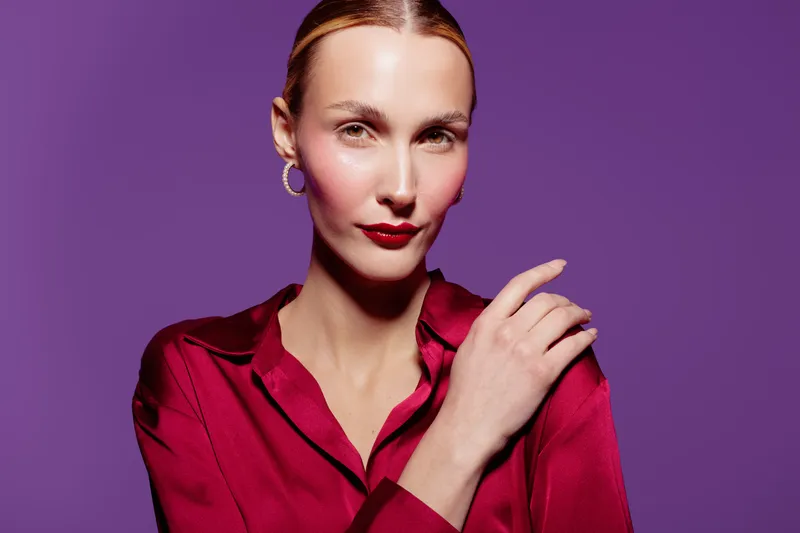 Confident woman in red satin blouse poses against purple background with hand near face. Studio portrait with elegant makeup and hoop earrings. Concept of fashion and beauty.
