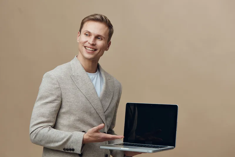 Smiling happy young man holding open laptop computer in hand posing isolated on beige background studio portrait. People lifestyle concept, casual style, positive emotions, technology, modern work env