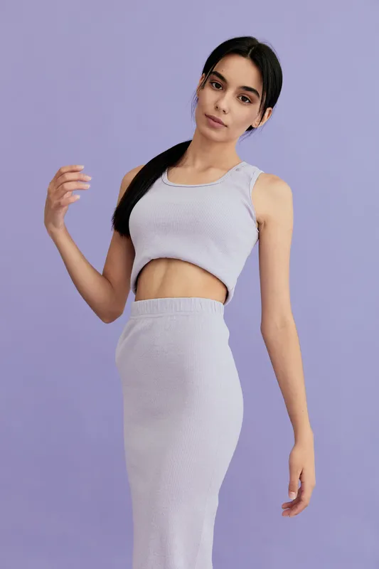 young woman in lavender two-piece outfit posing gracefully against a solid purple background, showcasing minimalist fashion and delicate features with natural makeup