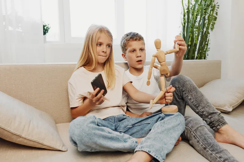 Two children sitting on a sofa in casual clothes, playing with a wooden mannequin and a smartphone in a bright living room. Concept of childhood, creativity, and home leisure time.