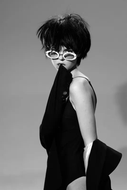 Stylish woman with short black hair wearing white sunglasses and black outfit in black and white portrait.