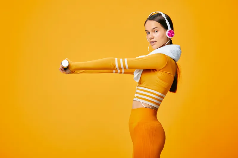 Fitness-minded woman in bright orange workout wear wears headphones and stretches with a dumbbell in a vivid studio, showcasing energy, balance and focus during a dynamic fitness session.