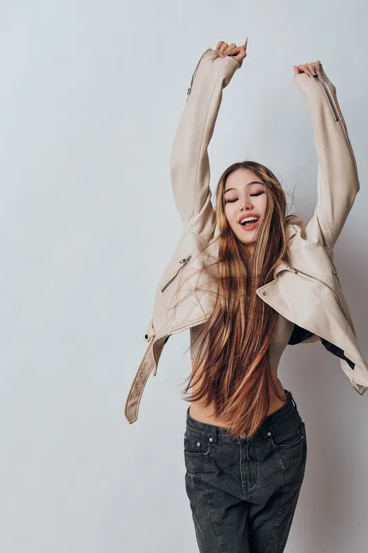Young woman with long hair and beige jacket stretching arms up and smiling with closed eyes on light grey background in casual clothes with relaxed and happy mood.