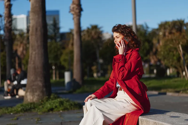 A woman sits on a concrete bench in a sunny park, talking on a mobile phone, wearing a red coat and white dress, depicting casual urban communication.