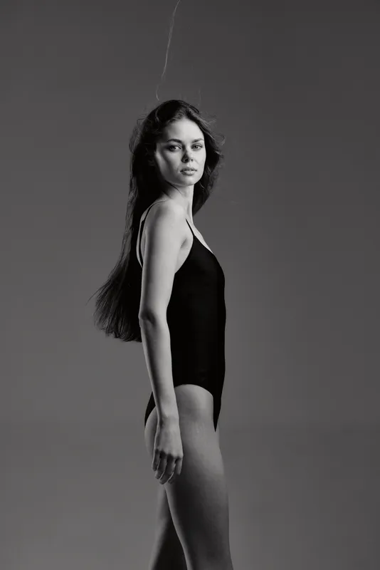 Black and white portrait of a woman in a black swimsuit with long hair, standing sideways against a plain background.