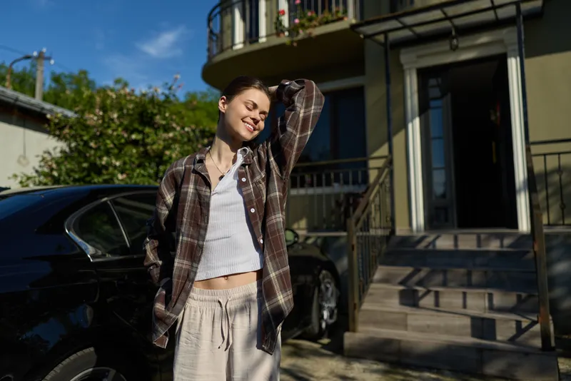 Young woman stands outside by a car, wearing a plaid shirt over a white cropped top and beige drawstring pants, smiling softly in bright afternoon sun