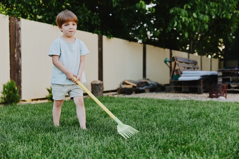 Young boy playing in the garden with a rake, enjoying outdoor activities on a sunny day, full of joy and innocence in a green lush environment.