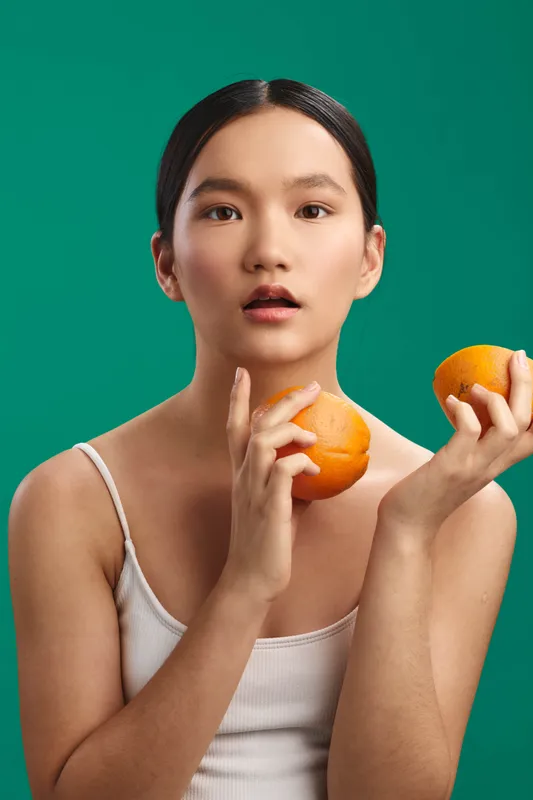 asian woman holds fresh oranges in a bright studio setup, wearing a white tank, calm expression amid a vibrant green background, creating a fresh, healthy lifestyle mood.