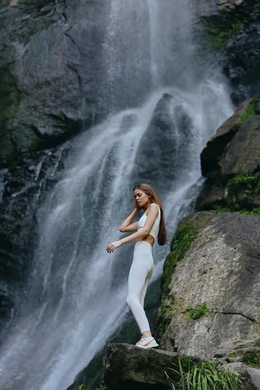 Young woman in white activewear standing on rock near a powerful waterfall surrounded by green vegetation and rocky cliffs.