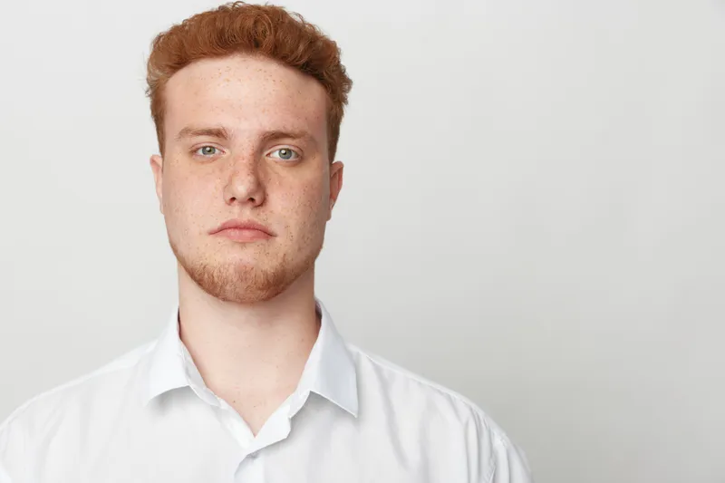 Portrait of a serious young man with red hair and freckles wearing a white shirt against a plain light background. Concept of confidence, youth, and natural appearance.